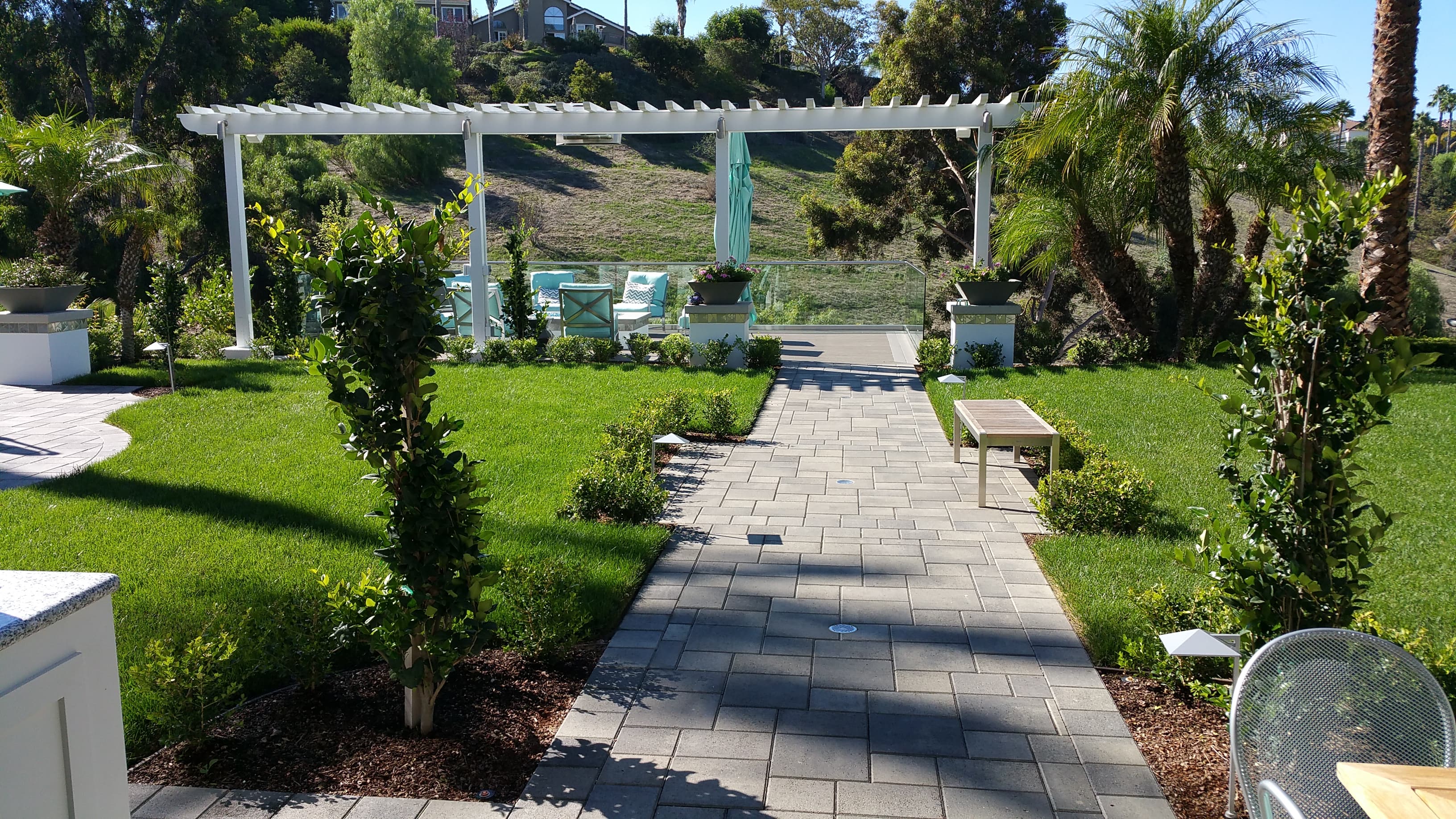 Paved walkway through a manicured lawn leading to a white pergola and lush green garden.
