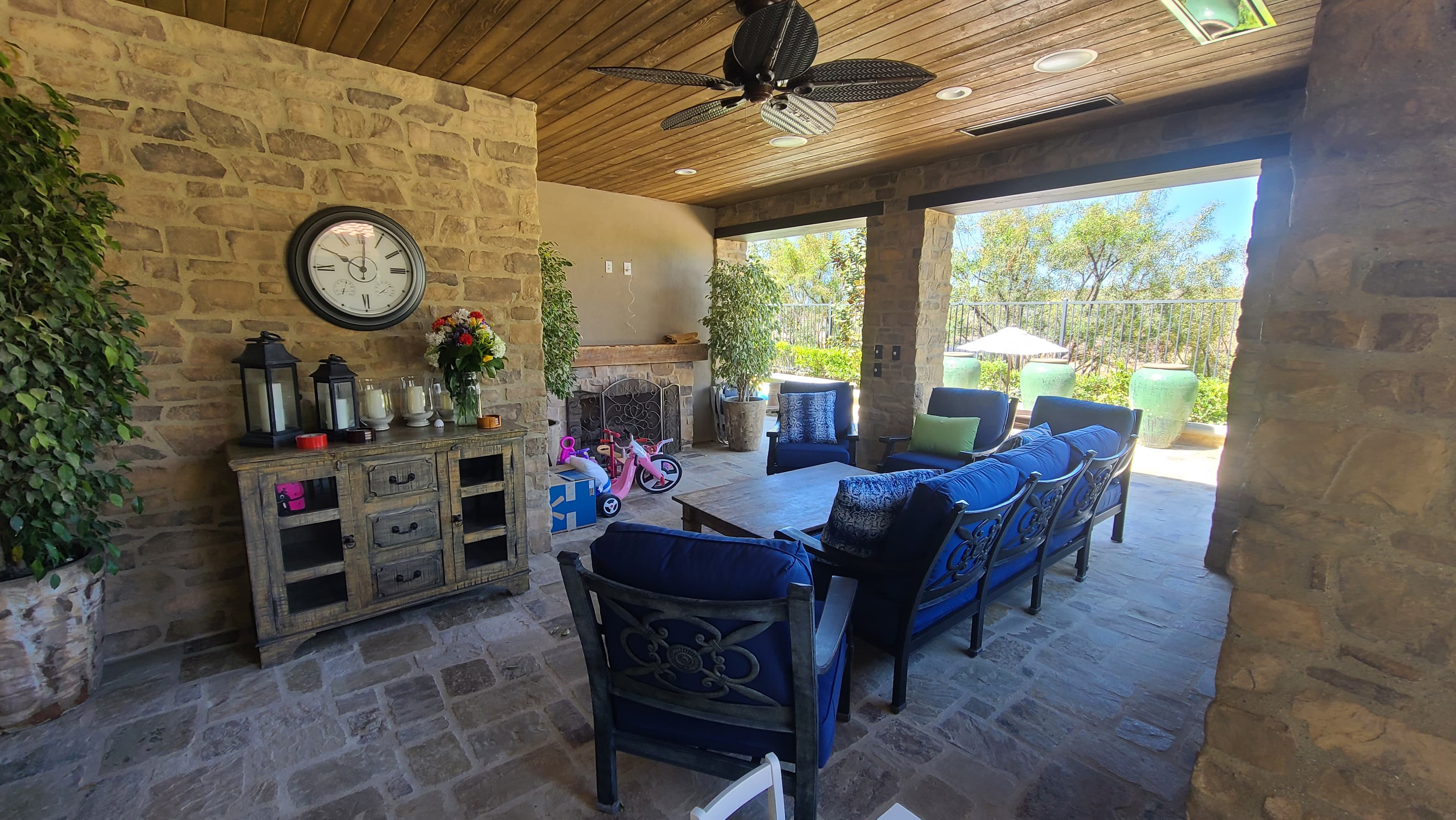 Covered stone patio featuring blue cushioned furniture, a wooden sideboard, and a ceiling fan.