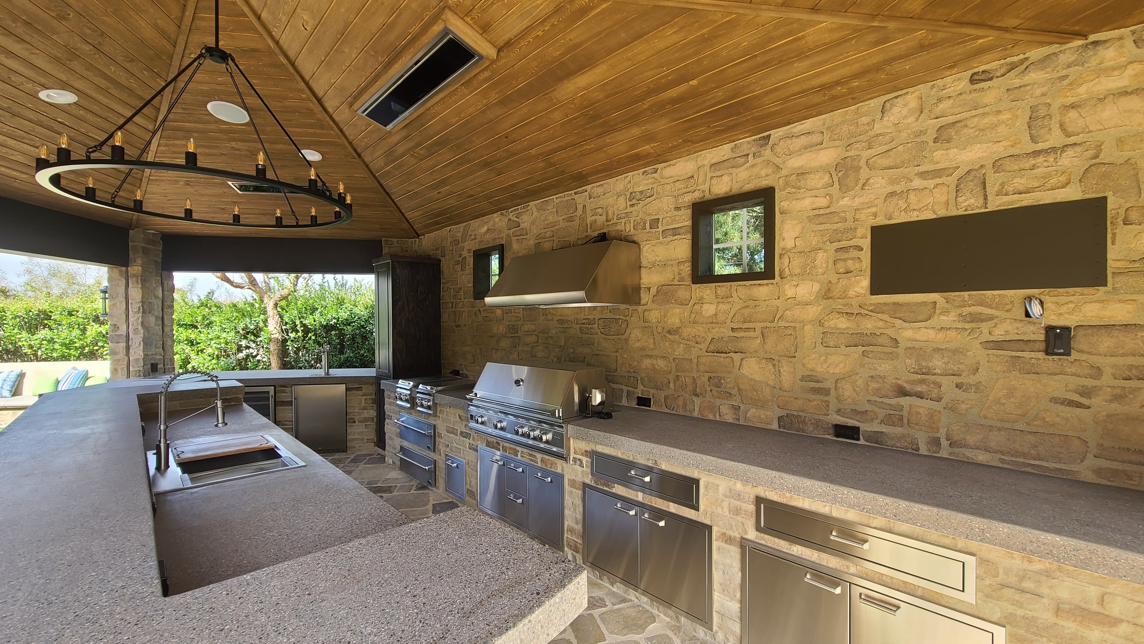 Stone-walled outdoor kitchen with stainless steel appliances, concrete countertops, and a wood-paneled ceiling.