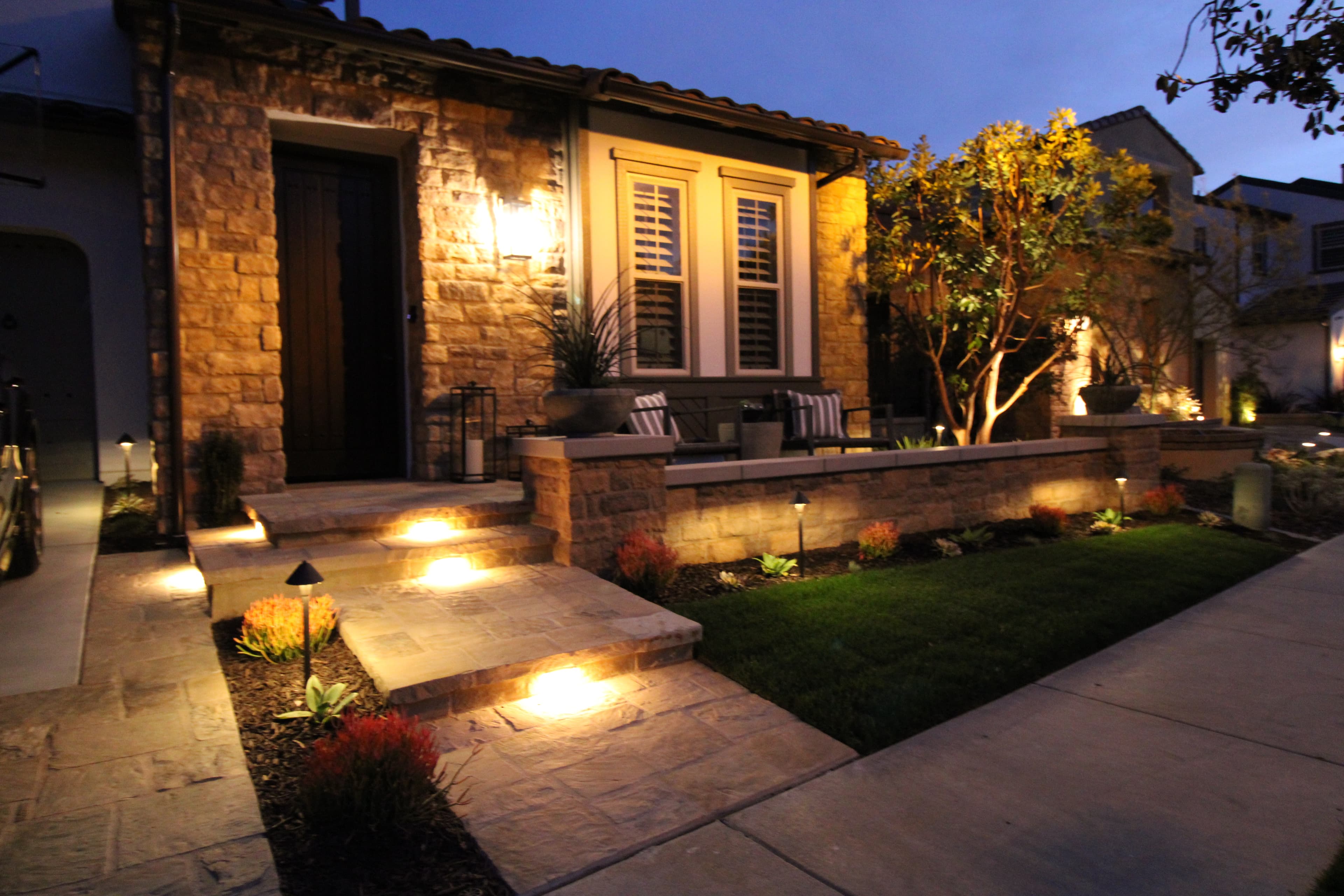 Warmly lit stone house exterior at night with glowing path lights and manicured landscaping.