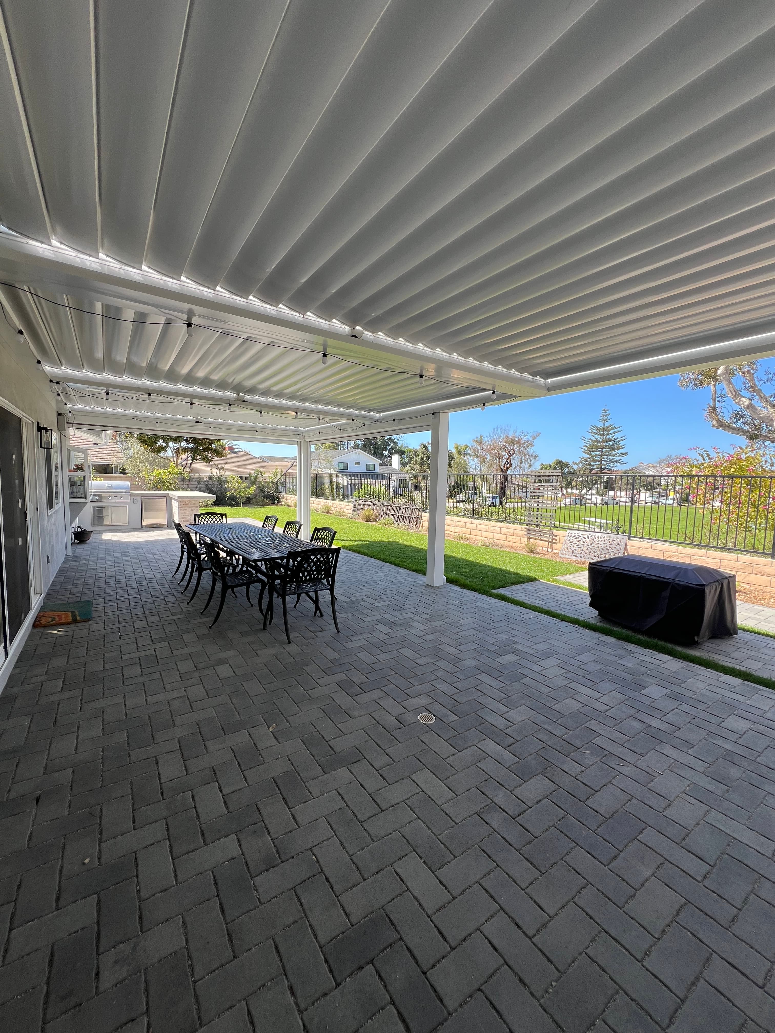 Modern outdoor patio featuring a white louvered pergola, grey brick pavers, and black dining furniture.