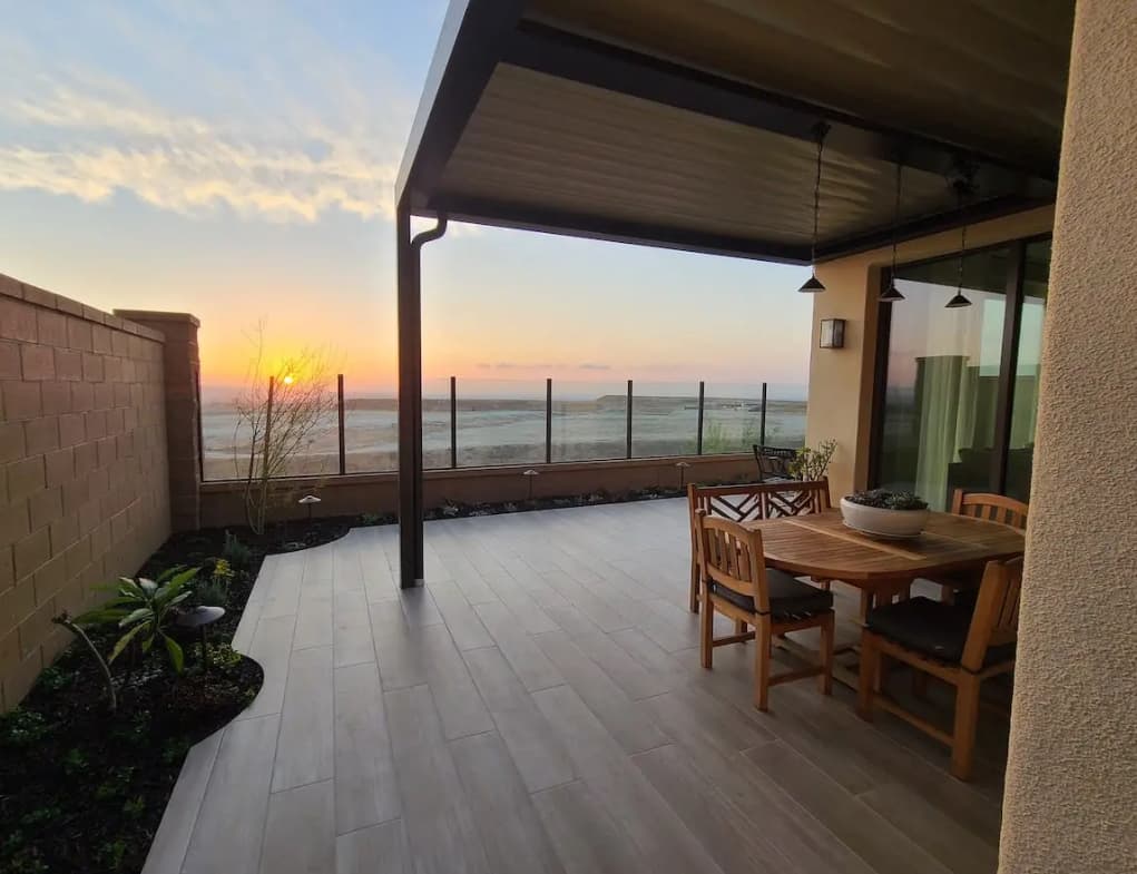 Modern tiled patio with wooden dining set and glass railing overlooking a scenic golden sunset.
