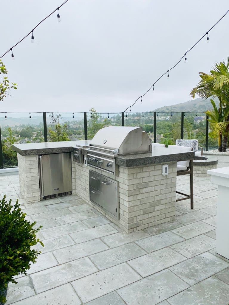 Modern outdoor kitchen with stainless steel grill and fridge on a stone patio overlooking hills.