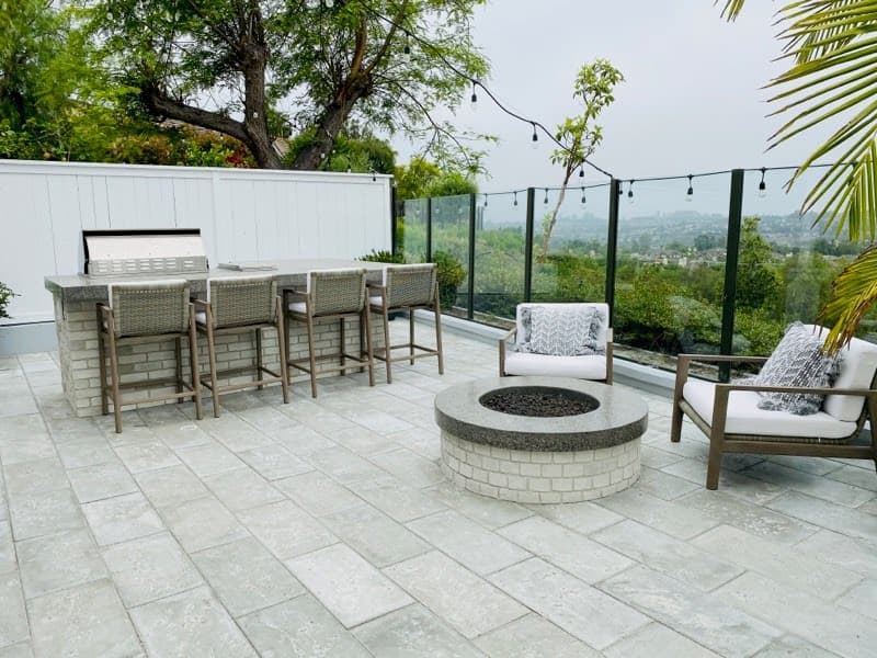 Modern stone patio featuring a fire pit, bar seating, and glass railing overlooking hills.